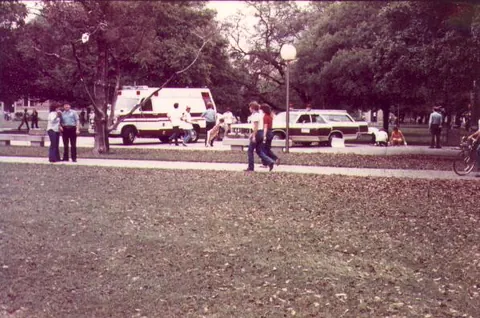 An ambulance and a patrol car parked on a tree-lined campus quad, with a few people gathered nearby in the distance.