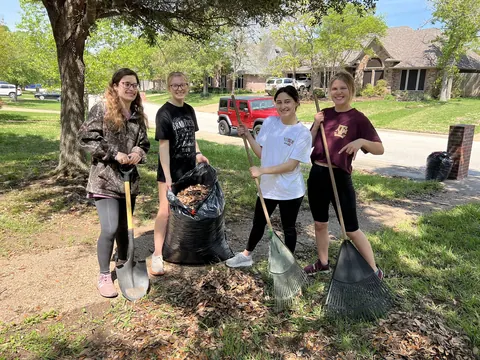 Four students holding rakes and shovels around a bag of yard waste on a sunny lawn.