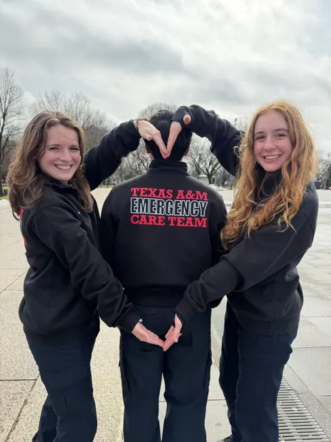 Two team members making a heart shape with their hands around the back of a jacket printed “Texas A&M Emergency Care Team”.