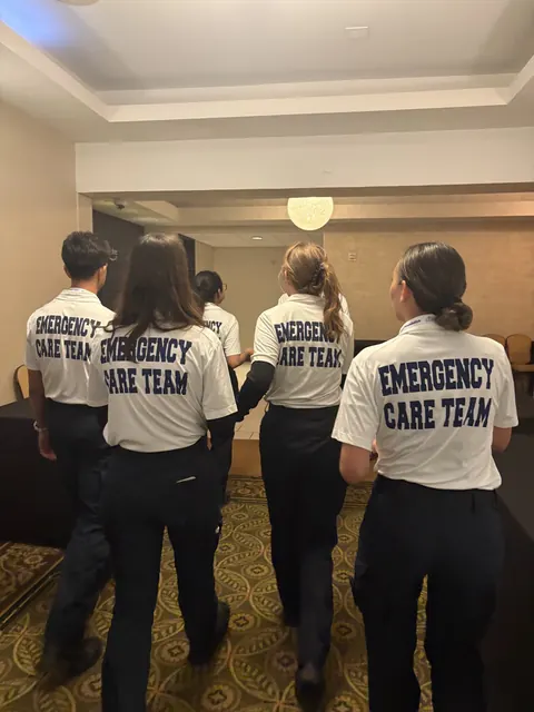 Four team members seen from behind in white shirts printed “Emergency Care Team”.